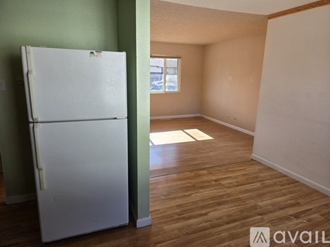 A white fridge stands in a room with wooden floors and a window.