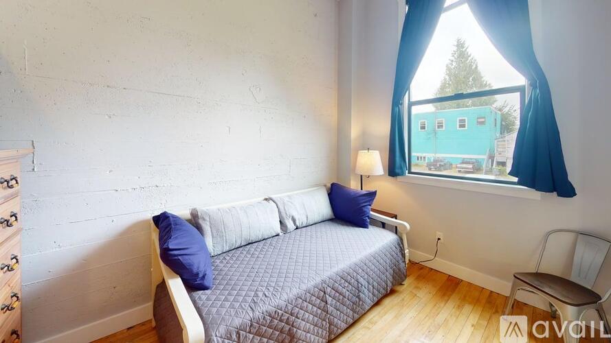 A bedroom with a bed, a window with a view of a blue building, and a wooden floor.
