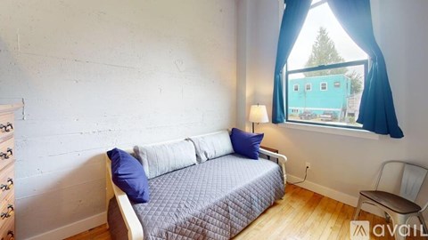 A bedroom with a bed, a window with a view of a blue building, and a wooden floor.