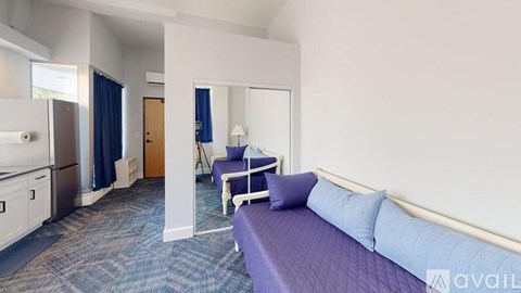 A bedroom with a purple bed and a kitchen area with a refrigerator and a sink.