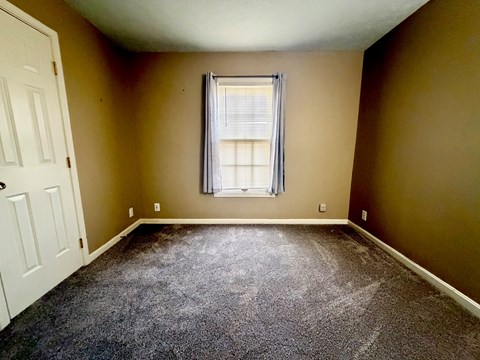 A room with a carpeted floor, a window with blinds, and a white door.