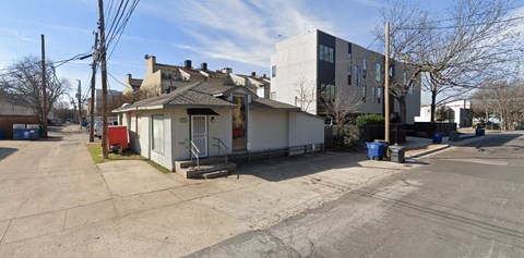 A small white house with a black roof is situated on a street with a blue trash can.