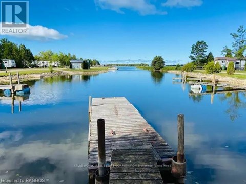 A wooden dock extends into a calm body of water with houses and trees in the background.