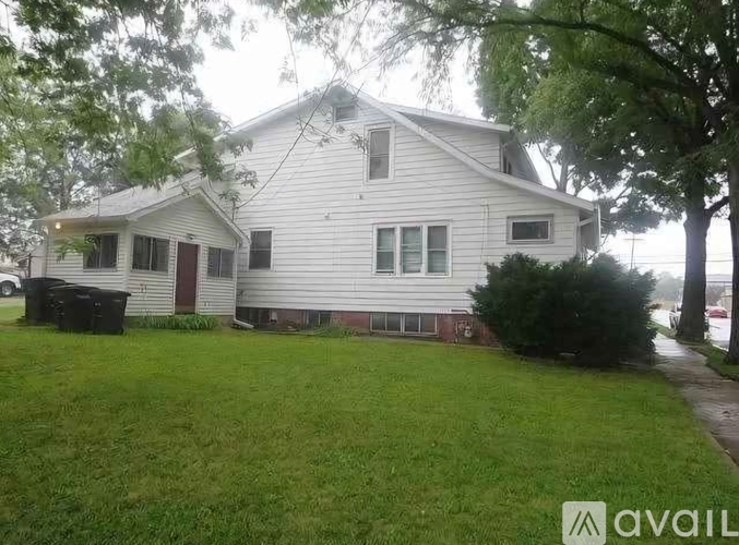 A house with a white siding and a red door is surrounded by a green lawn.