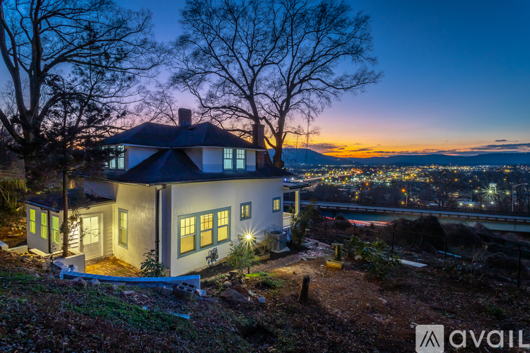 A house with a dark roof and white walls is surrounded by trees and has a view of a city at night.