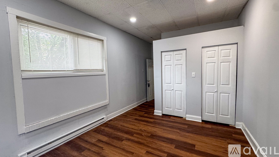 A room with wooden floors and white walls, featuring a window with blinds and two white doors.