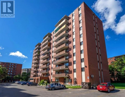 A tall red brick apartment building with a parking lot in front.