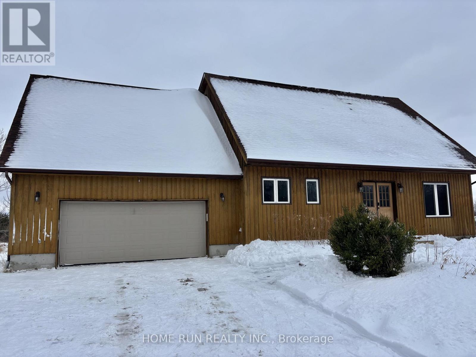 A house with a brown wooden exterior and a white garage door is surrounded by snow.