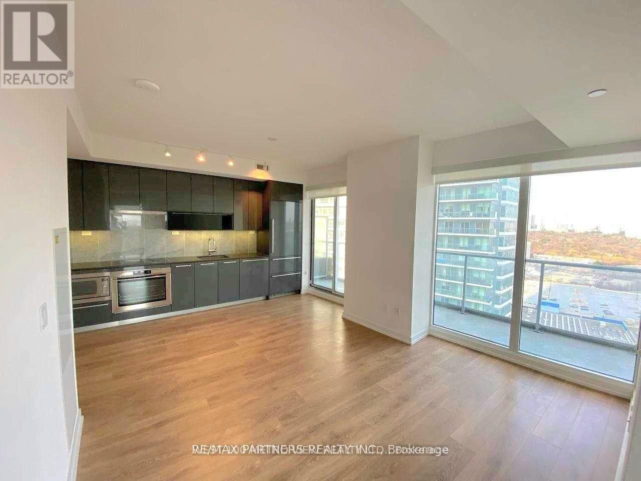 A spacious living room with hardwood floors and a large window overlooking a parking lot.