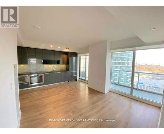 A spacious living room with hardwood floors and a large window overlooking a parking lot.