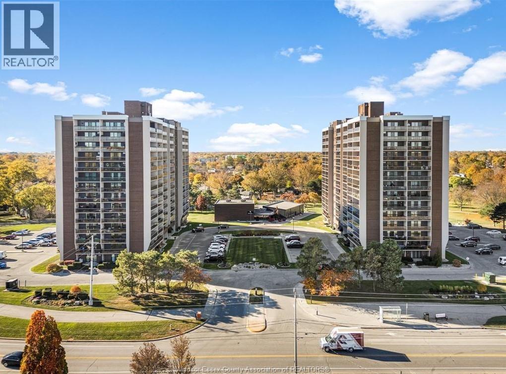 Two tall buildings are surrounded by a parking lot and trees.