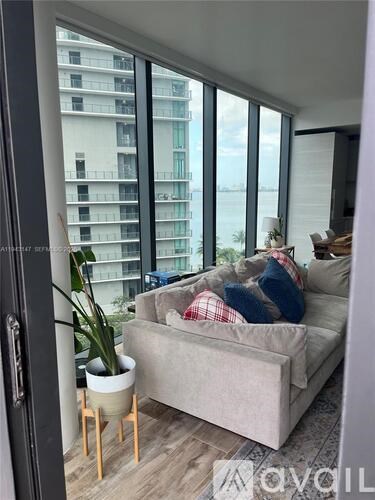 A living room with a couch, a potted plant, and a window overlooking a building.