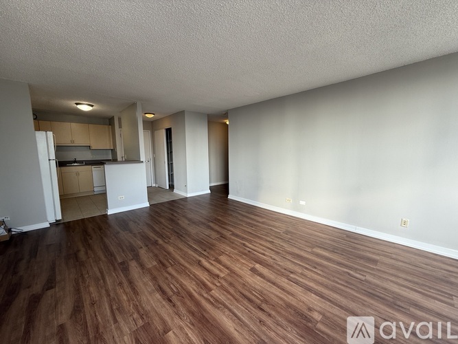 A room with wooden flooring and white walls, with a kitchen visible in the background.