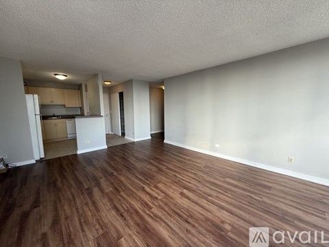 A room with wooden flooring and white walls, with a kitchen visible in the background.