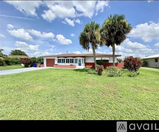 A house with a red roof and white walls is surrounded by a grassy lawn and palm trees.