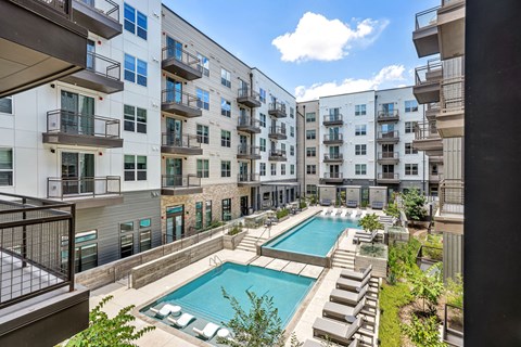 A swimming pool is surrounded by lounge chairs in a courtyard surrounded by apartment buildings.