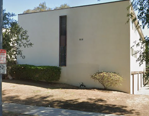 A small white building with a brown door and a small tree in front.