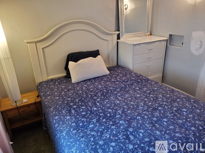 A bed with a blue patterned cover and a white pillow is in a room with a white dresser and mirror.