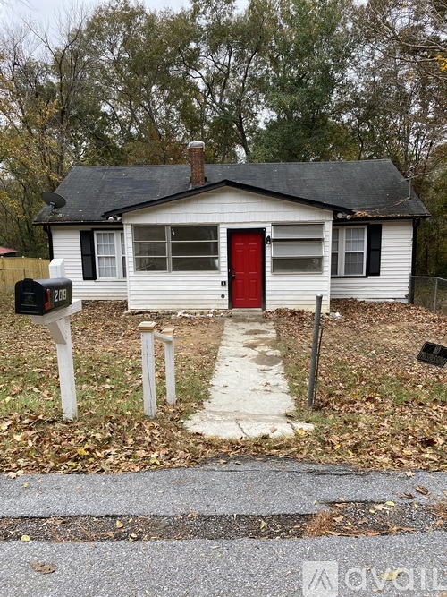 A small white house with a red door is surrounded by a grassy area and a mailbox is on the left.