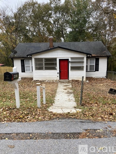 A small white house with a red door is surrounded by a grassy area and a mailbox is on the left.