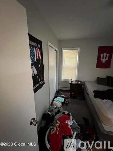 A bedroom with a bed, a window with blinds, a dresser with clothes, a poster on the wall, and a flag on the wall.