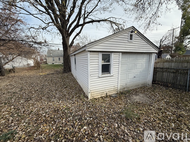 A small white shed with a garage door is surrounded by a gravel area with fallen leaves.