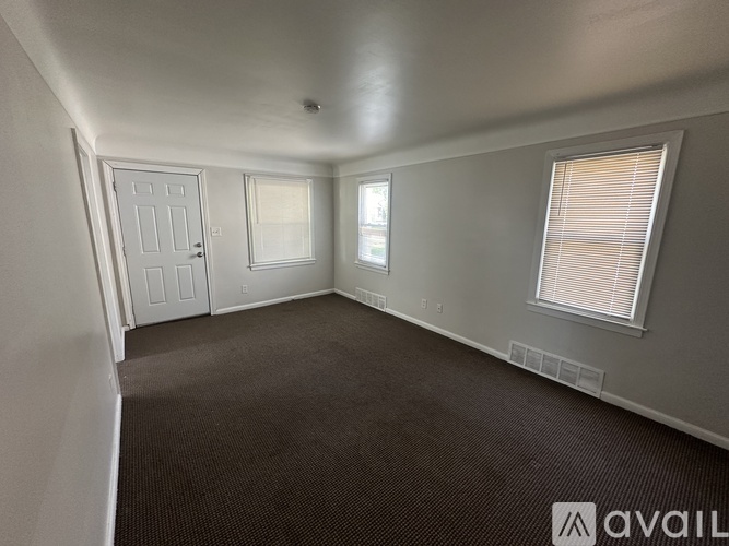 A room with a carpeted floor, a door, a window with blinds, and a wall-mounted shelf.