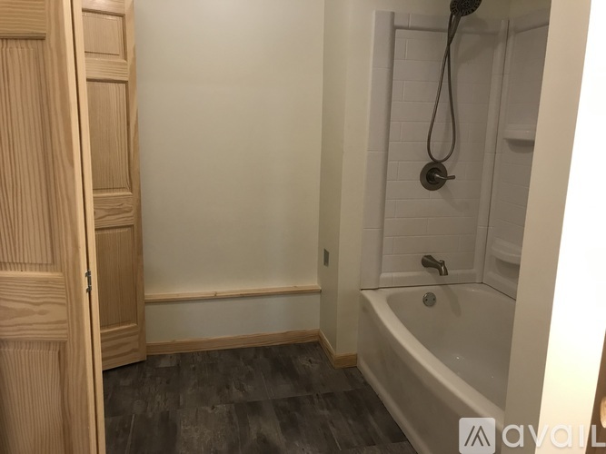A wooden door is on the left of a bathroom with a white tub and a shower head.