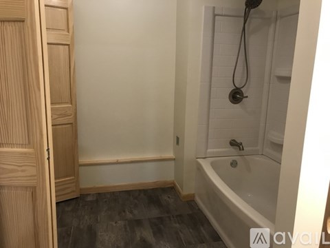 A wooden door is on the left of a bathroom with a white tub and a shower head.