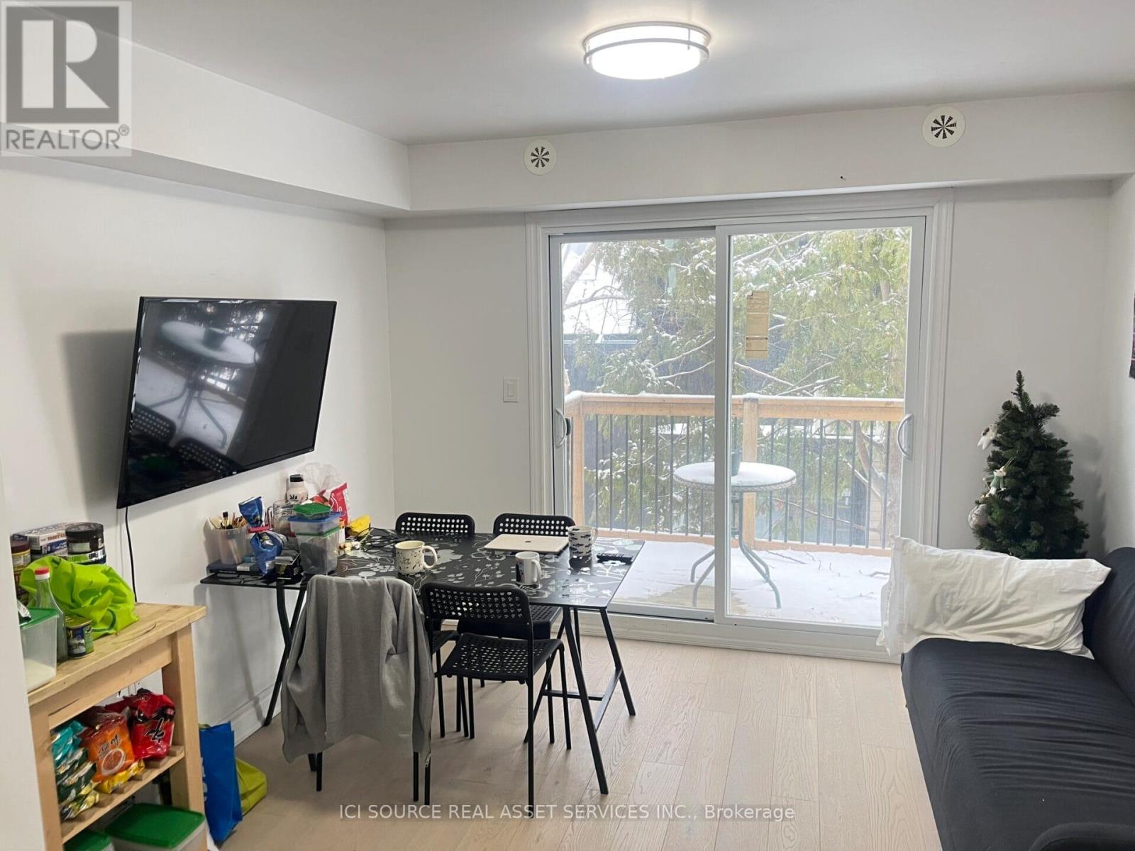 A living room with a grey couch, a television, a dining table with chairs, and a balcony with a view of trees.
