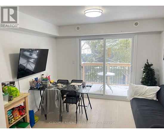 A living room with a grey couch, a television, a dining table with chairs, and a balcony with a view of trees.
