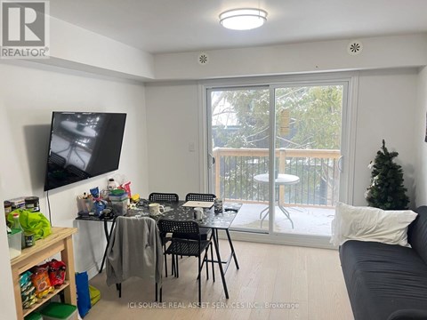A living room with a grey couch, a television, a dining table with chairs, and a balcony with a view of trees.