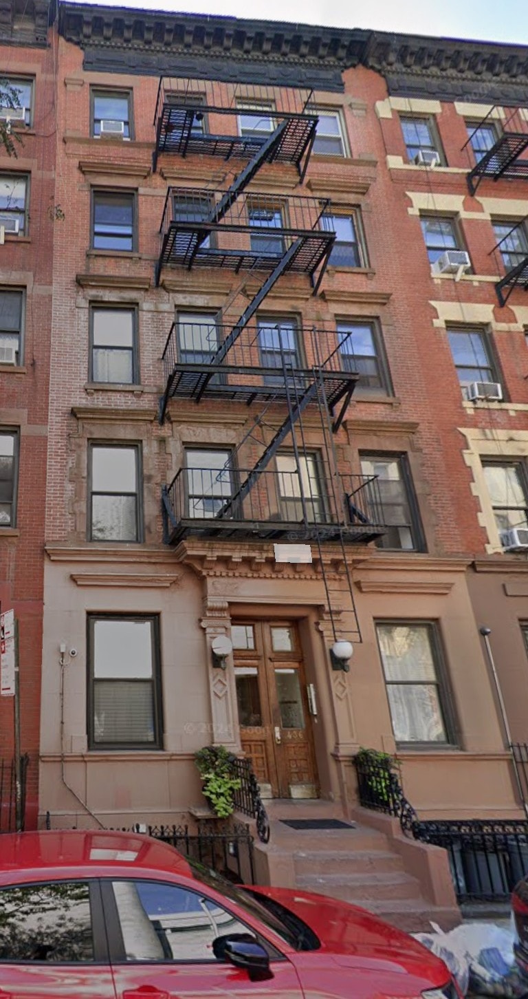 A red car is parked in front of a brown building with a black fire escape.