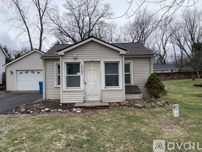A small house with a white door and windows is surrounded by a gravel driveway and a grassy area.