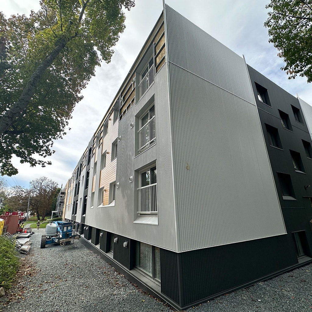 A modern building with a grey facade and black windows is surrounded by trees and a gravel path.