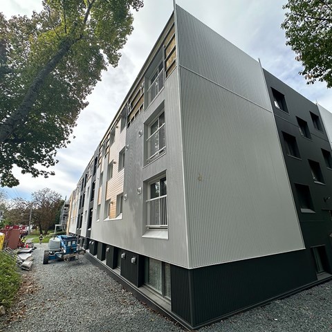 A modern building with a grey facade and black windows is surrounded by trees and a gravel path.