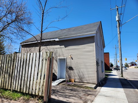 A small house with a grey roof and a white door is surrounded by a wooden fence.