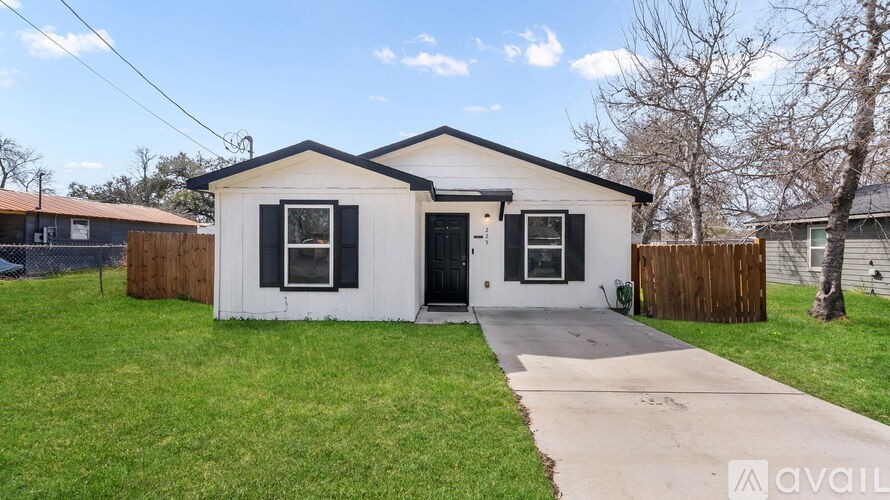 A small white house with a black door and windows is surrounded by a wooden fence and has two cars parked in front.