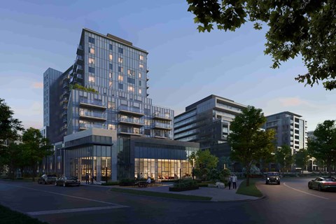 A modern multi-story building with a glass facade and balconies is surrounded by trees and has a parking lot in front.