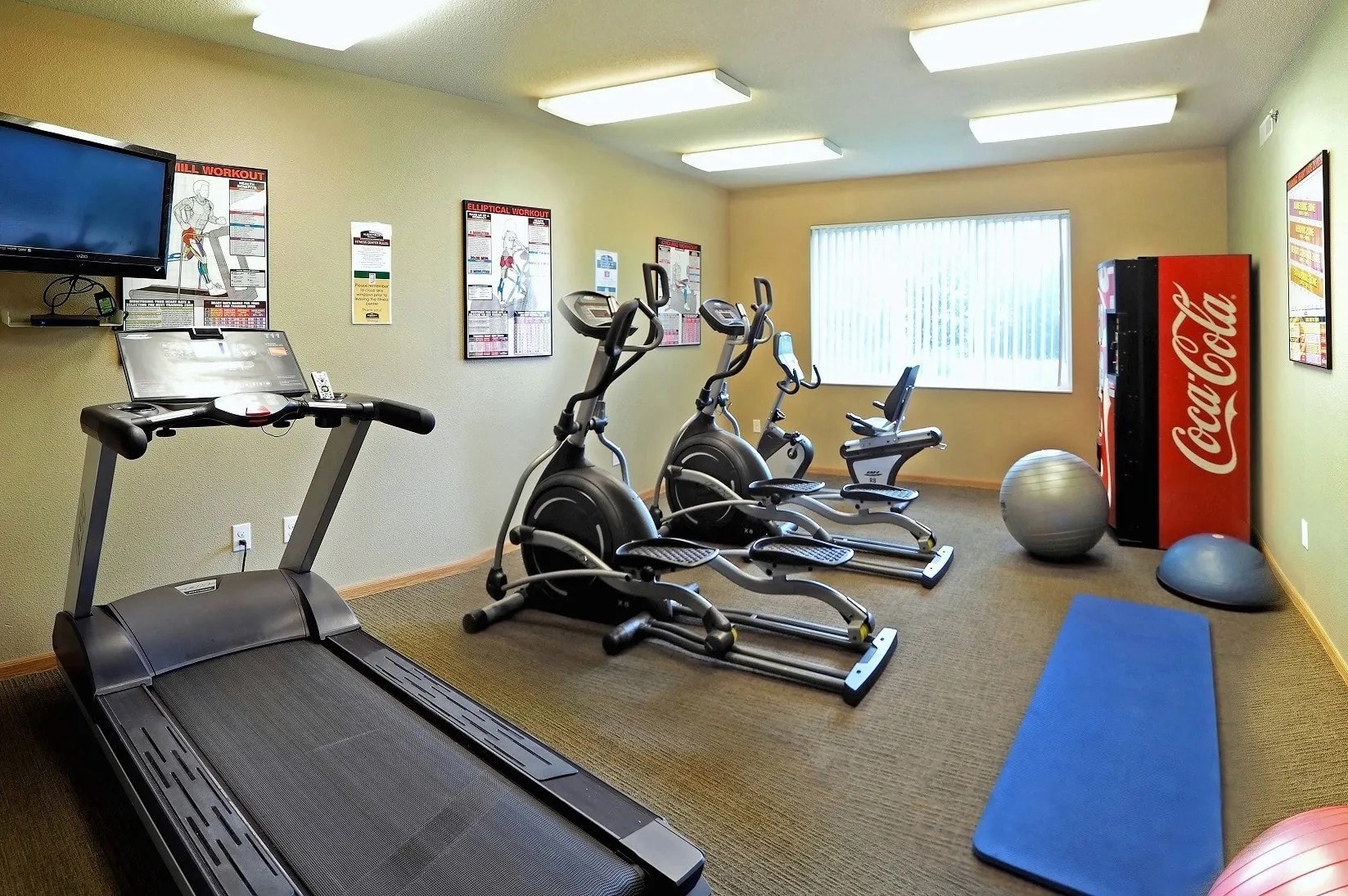 A Coca-Cola vending machine sits in a room with exercise equipment.