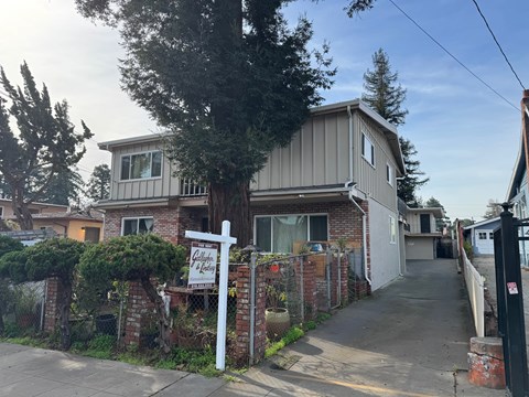A house with a sign that says "For Sale" is surrounded by a fence and trees.