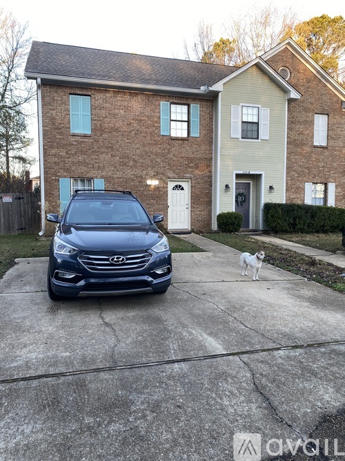 A blue car is parked in front of a house with a white dog standing on the concrete.