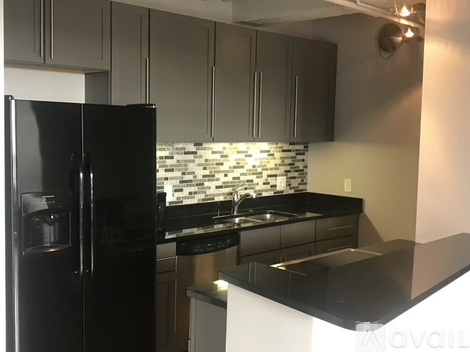 A black refrigerator stands in a kitchen with a black countertop and a backsplash of white and grey tiles.