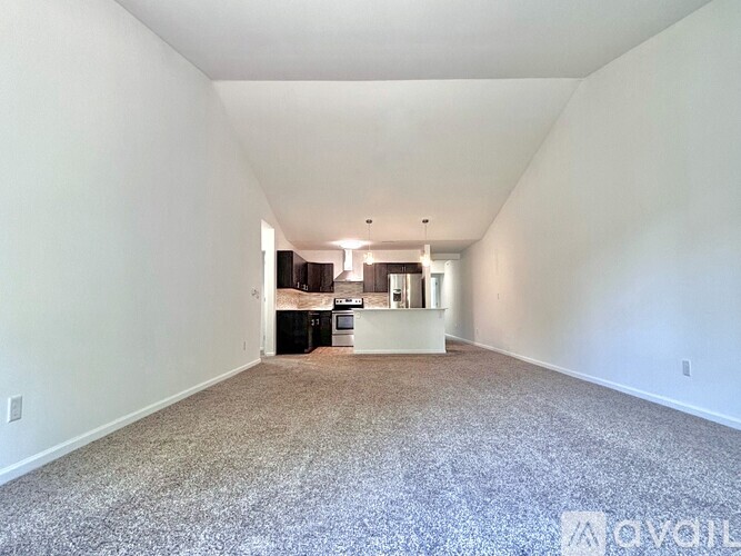 A large empty room with a carpeted floor and a kitchen area in the background.