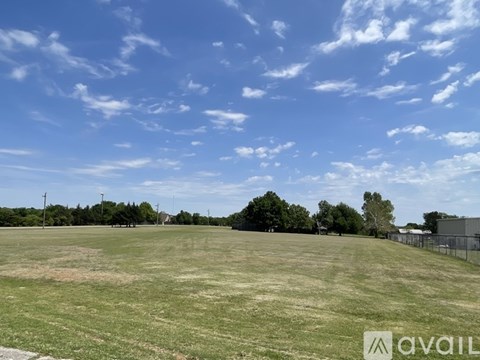 A large open field with a fence on the right side and trees in the distance under a blue sky with scattered clouds.