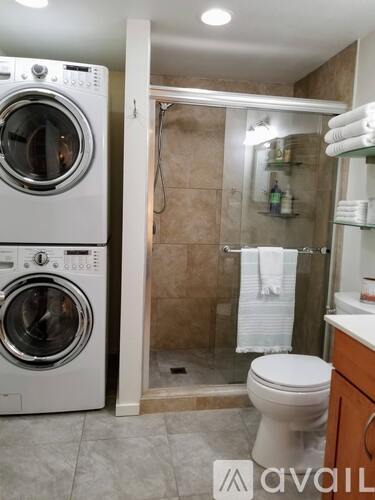 A white washing machine is in a bathroom with a toilet and a shower.