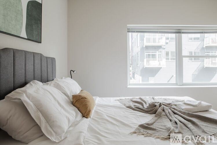 A bed with white sheets and a tan pillow is in front of a window with a view of buildings.
