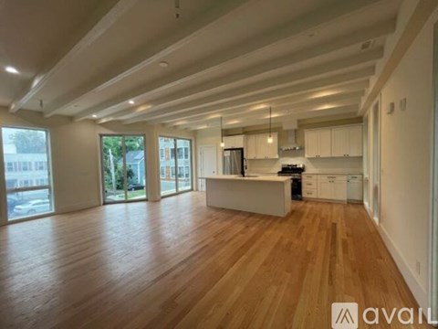 A spacious living room with wooden floors and a kitchen area in the background.