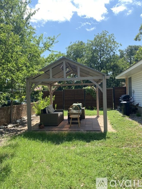 A wooden gazebo with a table and chairs is situated in a backyard.