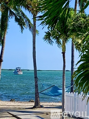 A boat is sailing in the distance behind a white fence and a few palm trees.